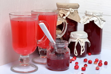 Homemade blanks. Cranberry jam in jars and glasses with cranberry juice. One can is open, it has a teaspoon. Several berries are scattered on the surface.