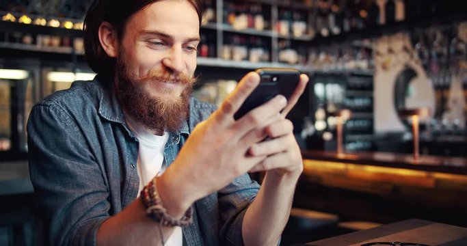 Stylish young male in casual outfit with bracelet on wrist chatting on smartphone, spending time in cafe, modern life