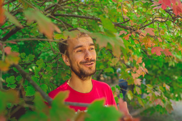 Naklejka na ściany i meble A man in the Park smiles standing behind the leaves. Portrait of a young man. Standing behind bushes and smiles