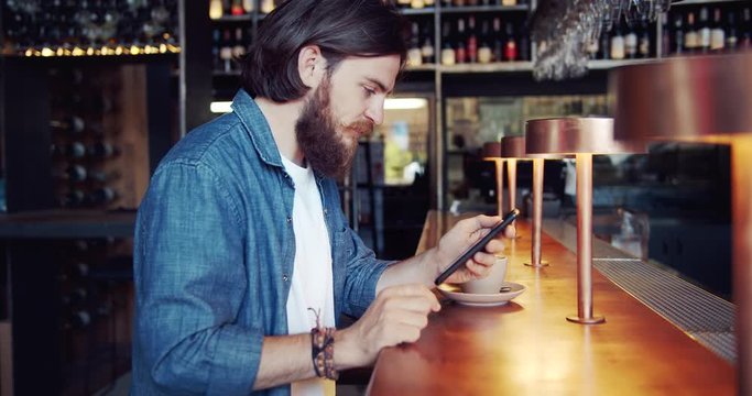 Attractive bearded male hipster in trendy clothing using mobile phone in bar and smiling, lifestyle