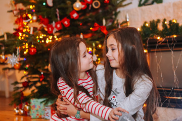 Obraz na płótnie Canvas Happy little kids under christmas tree at home. Girls play near christmas tree. Family with kids celebrate winter holidays in decorated living room with christmas lights. Xmas and New Year theme