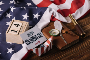 card with happy Columbus Day inscription near calendar, paper boat, nib, compass, telescope and notebook on wooden surface with American national flag