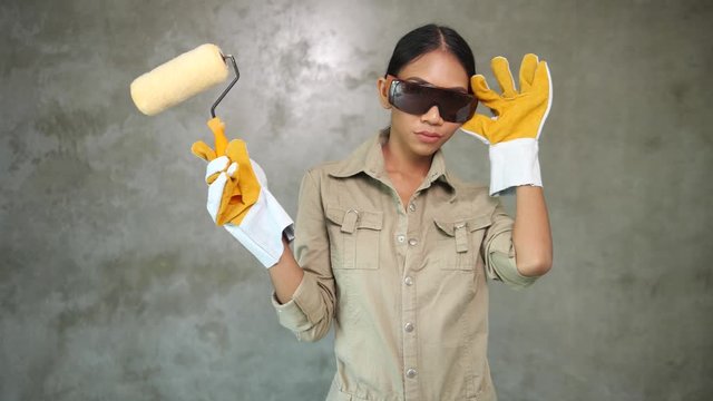Attractive young girl builder with paint roller in uniform, protective glasses and gloves smiling over concrete wall background
