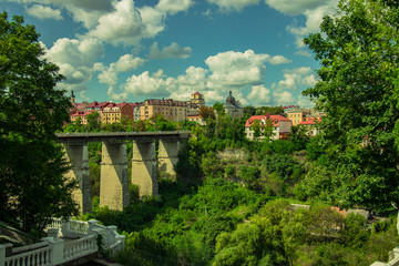 Fototapeta na wymiar travel poster concept photography of beautiful Ukrainian medieval city landmark view with colorful small living houses and high stone bridge across canyon in green trees foliage natural frame 