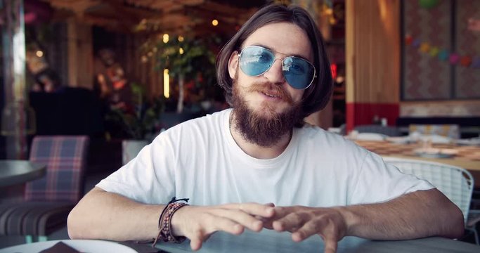 Young beaded hippie blogger in trendy eyewear recording video in fancy restaurant, sharing experience with community