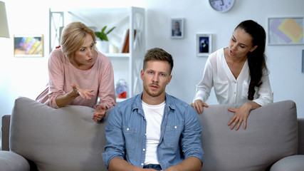 Irritated man sitting on sofa, listening wife and mother reproach, problem
