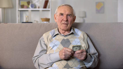 Aged male holding dollars sitting on sofa looking camera, low living standard