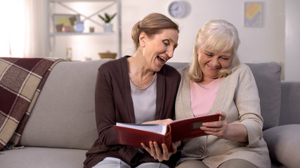 Positive aged ladies looking album photos sitting sofa at home, cheerful friends
