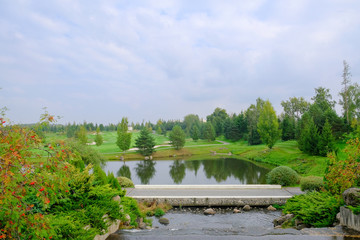 Landscape. Hills with green lawn and ornamental shrubs and trees lake and bridge. Beautiful garden