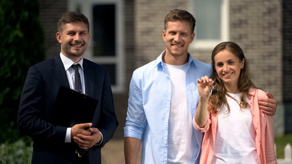 Smiling realtor with documents and happy couple with keys posing near house