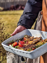 Man holding barbecued vegetables and steaks in white ceramic bowl. Autumn cooking in the garden.