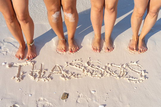 High Angle View Of Four Female Legs Barefoot Feet Standing On The Sand Beach By The Thassos Written On The Sand