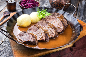 Traditioneller geschmorter deutscher Sauerbraten vom Rind mit Kartoffel Knödel und Blaukraut in würziger brauner Sauce als closeup in einer rustikalen schmiedeeisernen Pfanne