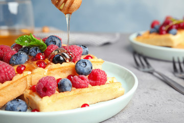 Pouring honey onto delicious waffles with fresh berries served on grey table