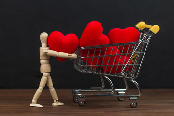 Wooden puppet loading red hearts in shopping cart / trolley on black background with copy space. Valentine's Day