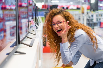 Red-haired girl with glasses standing at the counter in the electronics store choosing a new big TV to buy. Sales season at the Mall