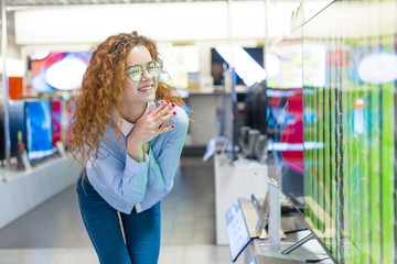 Red-haired girl with glasses standing at the counter in the store of electronic equipment choosing a new plasma to buy. Sales season at the Mall