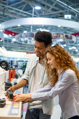 Young couple redhead girl with African man standing at the counter in the electronics store choosing a portable wireless music speaker to buy. Sales season at the Mall