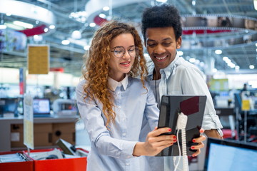 Young couple redhead girl in glasses with African man standing at the counter in the electronics store choosing a new tablet computer to buy. Sales season at the Mall
