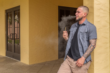 The young man leans on the wall vaping smoke while focused on the smoke surrounding him. Smoking...