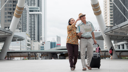 asian chinese senior tourist couple with black luggage walking together in city with map on hand...