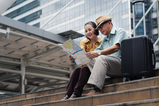 asian chinese senior tourist couple with black luggage sitting together on stairway urban with map on hand after having city sightseeing tour