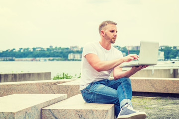 Fototapeta na wymiar Young American Man with beard, traveling, working in New York City, wearing white T shirt, blue jeans, sneakers, sitting on stone bench at park by river, hand holding laptop computer, reading, typing