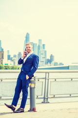 Young American Businessman with beard, talking on cell phone, traveling in New York City, wearing blue suit, white shirt, standing in business district with high buildings in Midtown of Manhattan..