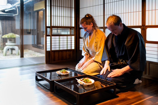 Couple in kimono bowing ojigi, seiza sitting at traditional Japanese home room by food plates on table with shoji sliding paper doors looking garden with stone lantern