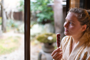 Traditional Japanese home or ryokan restaurant with young woman sitting by window in kimono or yukata looking at garden eating anko dango