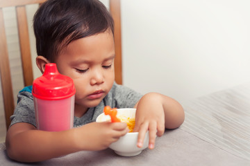 A toddler sitting at the dinner table without a high chair and playing with his food while holding a spoon.