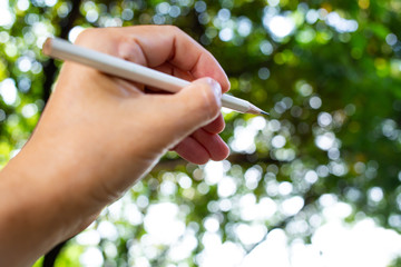 Woman's left Hand holding white pencil, Writing letter in bokeh green garden background, Close up & Macro shot, Selective focus, Image, Communication, Stationery concept