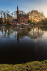 Fototapeta na wymiar Heritage of brick building reflects into lake with dry tree background at Minnewater park during sunrise, Bruges, Belgium