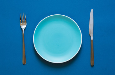 Ready to order. High angle shot of empty color plate, fork, knife lying against blue background.