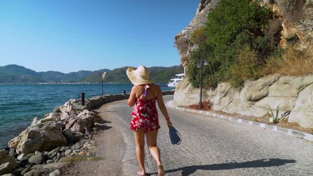 This shot depicts a panoramic view of a curvy woman with a straw hat and jelly shoes walking by the seaside in Turkey, Marmaris.