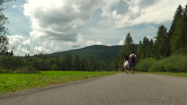 Back of couple walking on paved road in forest area with clouds on sunny day