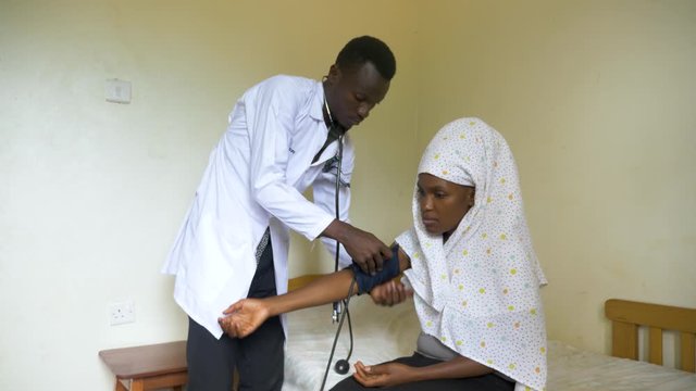 Wide shot of a young male African doctor checking the blood pressure of a Muslim African lady in a small health clinic in rural Africa.