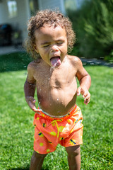 Cute little boy drinking from the sprinklers in the backyard on a hot summertime day. African American boy playing in the outdoors trying to stay cool on a warm summer day