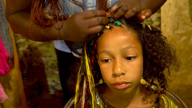 African hairdresser plaits braids with multicolored artificial strands for mixed Jamaica ethnicity little girl. Portrait of a girl with curly long hair