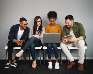 Multiethnic friends sitting on chair obsessed with modern devices looking at laptop while waiting...