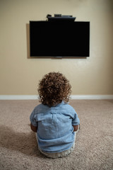 Rear view of a young child watching Television while sitting on the floor of his home. Selective focus on the back of the curly-haired diverse little boy.