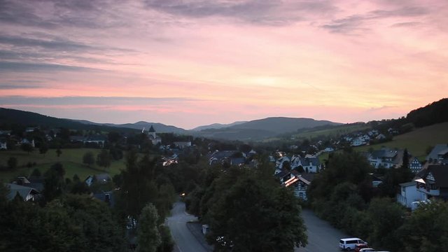 Wide panoramic pan over the mountainous spa village of Grafschaft in the winter sports region of Sauerland, Germany, during sunset with a colourful purple and orange lit cloudy sky