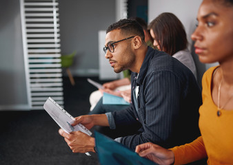 Side view of a confident young african american man wearing eyeglasses holding resume in hand waiting for job interview with other candidates
