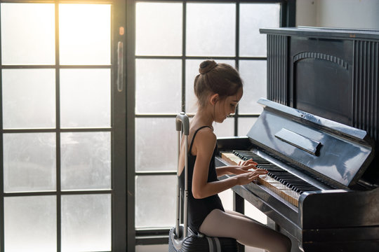 Cute beautiful young little girl ballet dancer in black leotard sitting on travel luggage and playing piano in dance studio.