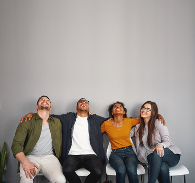 Group Of Multiracial Young Friends Sitting On Chair With Their Arms Around On Each Other's Shoulder Having Fun And Looking Up At Copy Space