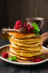 Stack of homemade little pancakes with honey, fresh raspberries and red currants on an old wooden background. Selective focus.