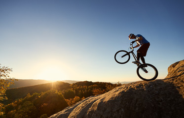 Fototapeta na wymiar Silhouette of cyclist balancing on back wheel on trial bicycle. Courageous sportsman biker making acrobatic trick on top of rocky mountain at sunset. Concept of extreme sport active lifestyle