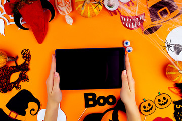 Female hands are holding tablet on orange background. Little pumpkins, photo props, masks, spiderwebs, decor for celebration are scattered in circle of canvas. Party accessories. Happy halloween.