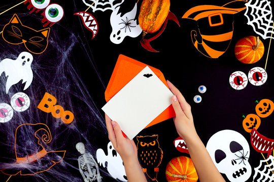 Female hands are holding envelope with blank sheet  for invitation on black background. Little pumpkins, photo props, decor for celebration are scattered on canvas. Party accessories. Happy halloween