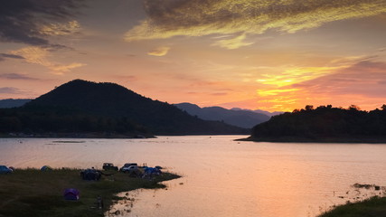 Fototapeta na wymiar Lake view evening of many camping tents on the bank around with the hills and yellow sun light with cloudy sky background, sunset at Kaengkrachan National Park, Phetchaburi, Thailand.